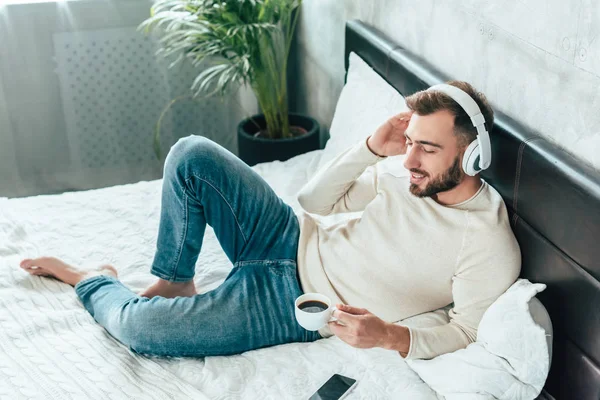 Vista aérea del hombre feliz escuchando música en auriculares mientras sostiene la taza de café en la cama - foto de stock