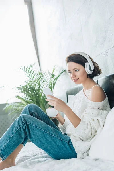 Mulher morena alegre segurando copo e usando smartphone enquanto ouve música em fones de ouvido — Fotografia de Stock