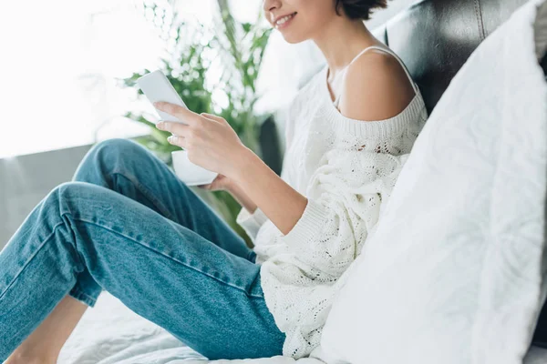 Vista ritagliata di donna felice in possesso di tazza e utilizzando smartphone in camera da letto — Foto stock