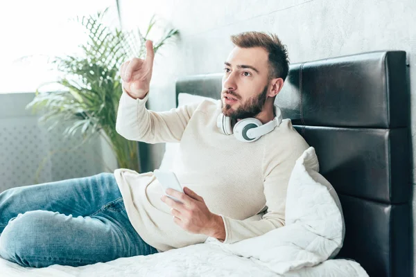 Schöner bärtiger Mann mit Kopfhörer, Smartphone in der Hand und gestikulierend im Schlafzimmer — Stockfoto