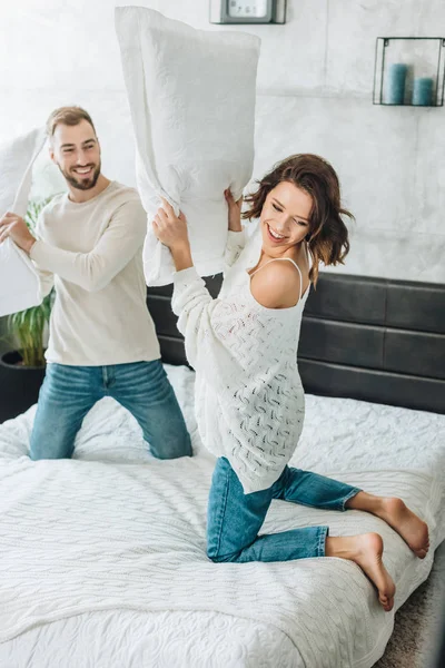 Happy bearded man having pillow fight with attractive woman on bed — Stock Photo