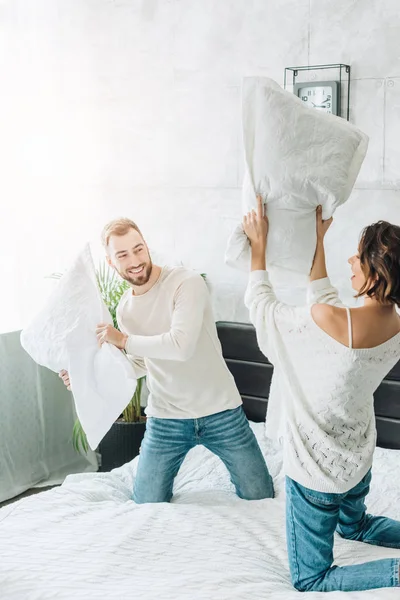 Happy bearded man having pillow fight with cheerful woman on bed — Stock Photo