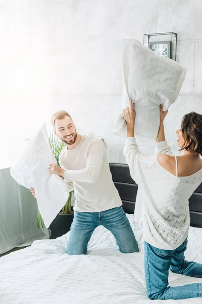 Heureux homme barbu ayant bataille d'oreiller avec femme gaie sur le lit — Photo de stock