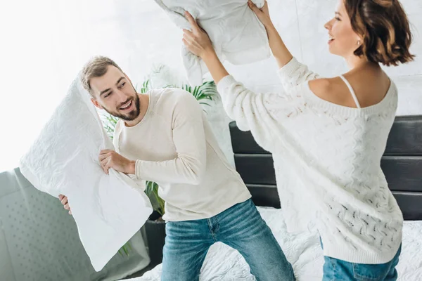 Enfoque selectivo de hombre barbudo feliz teniendo pelea de almohadas con mujer alegre en la cama - foto de stock