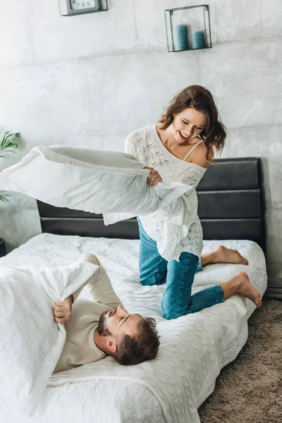 Femme gaie ayant bataille d'oreiller avec l'homme barbu heureux sur le lit — Photo de stock