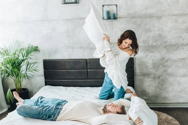Веселая женщина, дерущаяся на подушках с красивым бородатым мужчиной, лежащим на кровати — стоковое фото