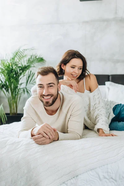 Homem barbudo alegre com as mãos apertadas olhando para a câmera com mulher atraente enquanto deitado na cama — Fotografia de Stock