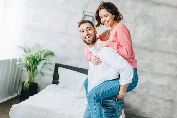 Feliz barbudo hombre piggybacking atractiva morena mujer en el dormitorio - foto de stock