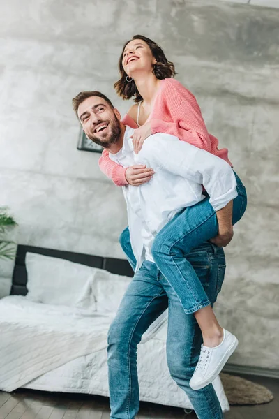 Vista de ángulo bajo de hombre barbudo feliz piggybacking mujer morena en el dormitorio - foto de stock