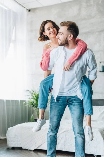 Cheerful bearded man piggybacking happy brunette woman at home — Stock Photo