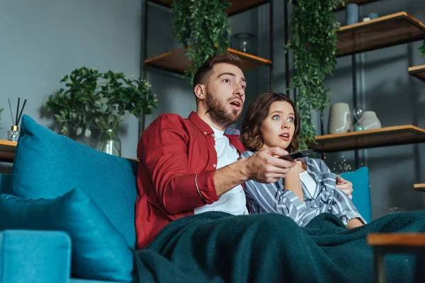 Vista de ángulo bajo del hombre sorprendido sosteniendo el mando a distancia mientras ve la película con la mujer en casa - foto de stock