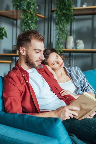 Enfoque selectivo de hombre feliz libro de lectura cerca de la mujer dormida en casa - foto de stock