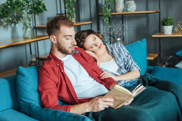 Guapo barbudo hombre lectura libro cerca de dormir mujer en sala de estar - foto de stock