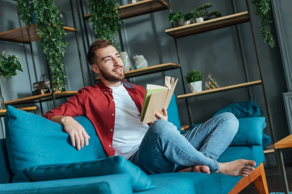 Schöner bärtiger Mann sitzt auf Sofa und hält Buch im Wohnzimmer — Stockfoto