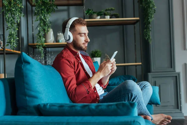 Selective focus of handsome bearded man listening music in headphones and using smartphone in living room — Stock Photo