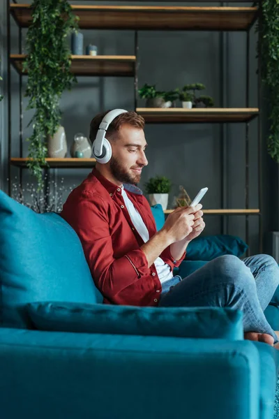 Selektiver Fokus eines fröhlichen bärtigen Mannes, der Musik über Kopfhörer hört und das Smartphone im Wohnzimmer benutzt — Stockfoto