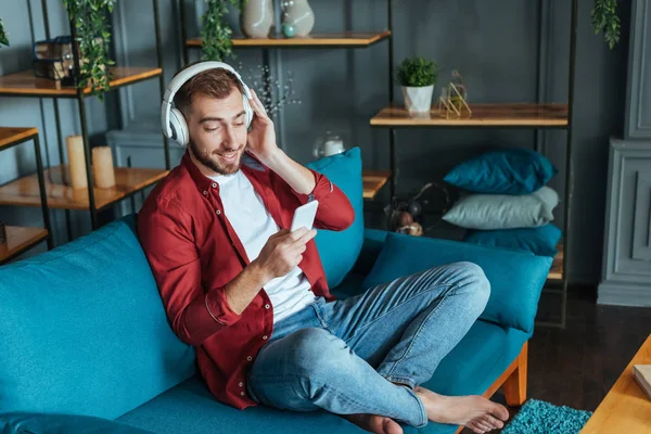 Glücklicher bärtiger Mann, der Musik über Kopfhörer hört und sein Smartphone im Wohnzimmer benutzt — Stockfoto