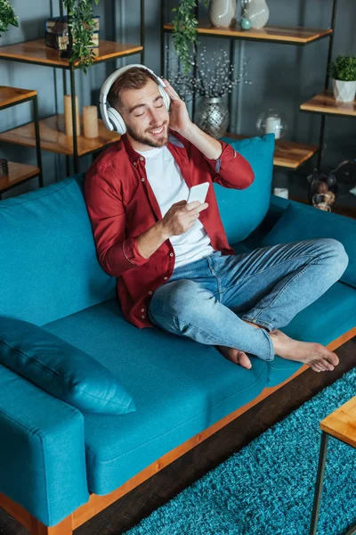 Vista aérea del hombre barbudo feliz escuchando música en los auriculares y utilizando el teléfono inteligente en la sala de estar - foto de stock