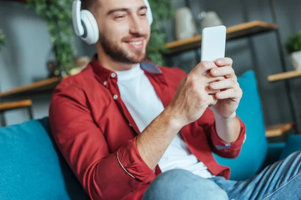 Foco seletivo de homem barbudo feliz ouvindo música em fones de ouvido e usando smartphone na sala de estar — Fotografia de Stock