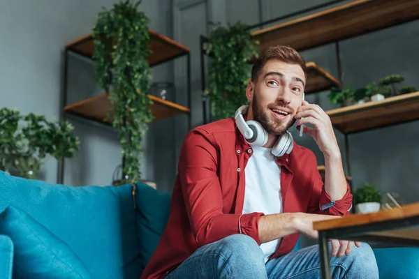 Vista de bajo ángulo del hombre barbudo feliz en auriculares hablando en el teléfono inteligente en la sala de estar - foto de stock