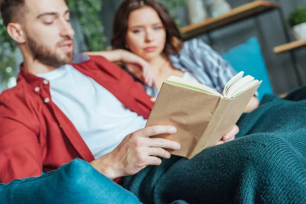 Enfoque selectivo de hombre libro de lectura cerca de mujer morena en casa - foto de stock