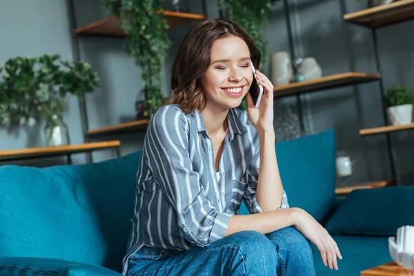 Mujer alegre con los ojos cerrados hablando en teléfono inteligente en la sala de estar - foto de stock