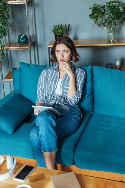 Mujer morena pensativa sosteniendo tableta digital mientras está sentado en la sala de estar - foto de stock