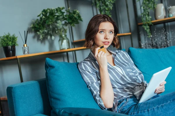 Mujer pensativa sosteniendo tableta digital y tarjeta de crédito en la sala de estar — Stock Photo
