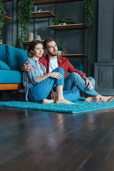 Blick in den niedrigen Winkel: glücklicher Mann umarmt attraktive Frau, während er auf Teppich im Wohnzimmer sitzt — Stockfoto