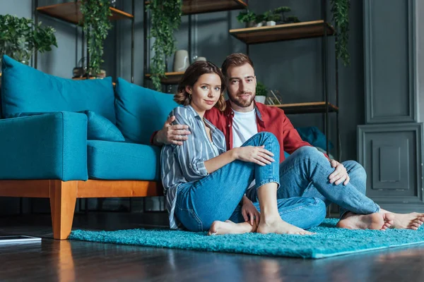 Low angle view of cheerful man hugging attractive woman while sitting on carpet at home — Stock Photo