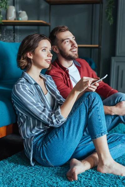 Attractive woman holding smartphone near happy man while sitting on carpet — Stock Photo