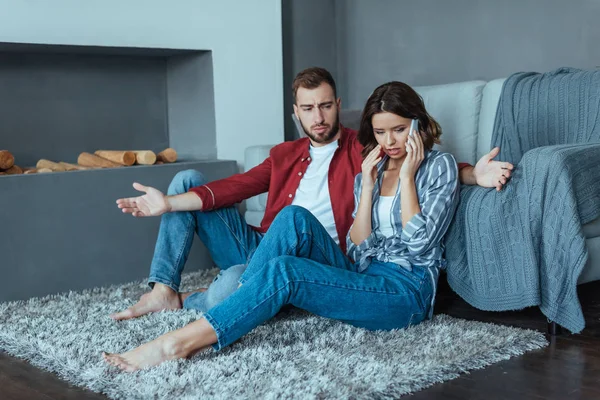 Upset man looking at attractive woman talking on smartphone while sitting on carpet — Stock Photo