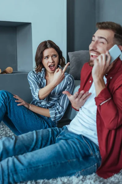 Selective focus of emotional woman showing middle finger while looking at happy man talking on smartphone — Stock Photo