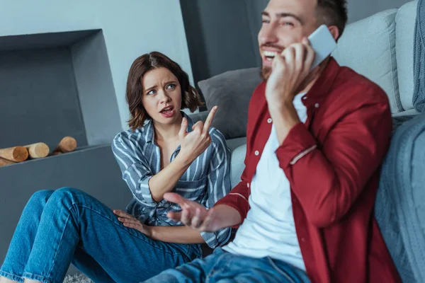 Messa a fuoco selettiva di donna sconvolta mostrando dito medio mentre guardando l'uomo felice che parla su smartphone — Foto stock