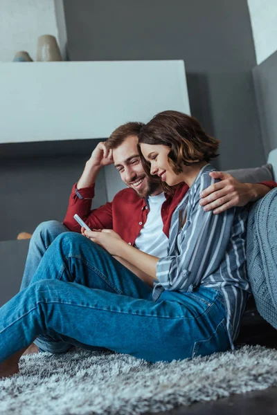 Baixo ângulo de visão do homem feliz e mulher olhando para o smartphone enquanto sentado no tapete — Fotografia de Stock