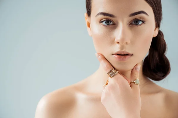 Junge Frau mit glänzendem Make-up und goldenen Ringen, die Gesicht isoliert auf grau berühren — Stockfoto