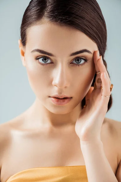 Mujer joven con labios brillantes, sombra de ojos dorada y anillos en los dedos mirando a la cámara aislada en gris - foto de stock