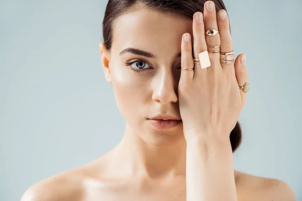 Giovane donna nuda con trucco lucido e anelli dorati che si nascondono dietro la mano isolata sul grigio — Foto stock