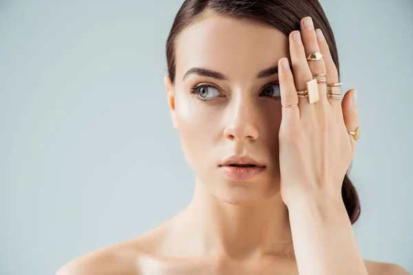 Mujer desnuda joven con maquillaje brillante y anillos de oro escondiendo la cara detrás de la mano y mirando hacia otro lado aislado en gris - foto de stock