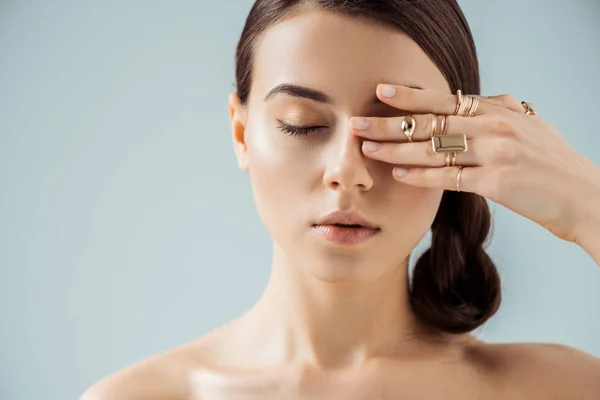 Junge nackte Frau mit glänzendem Make-up und goldenen Ringen versteckt Auge hinter Hand isoliert auf grau — Stockfoto