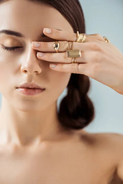 Mujer desnuda joven con maquillaje brillante y ojos cerrados en anillos dorados ocultando el ojo detrás de la mano aislado en gris - foto de stock