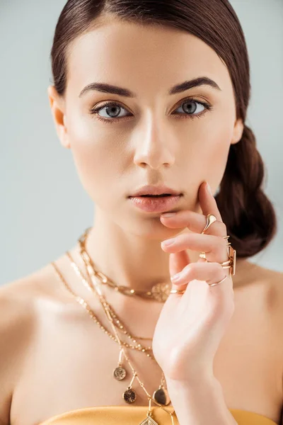 Mujer joven con maquillaje brillante en collares dorados y anillos mirando a la cámara aislada en gris - foto de stock