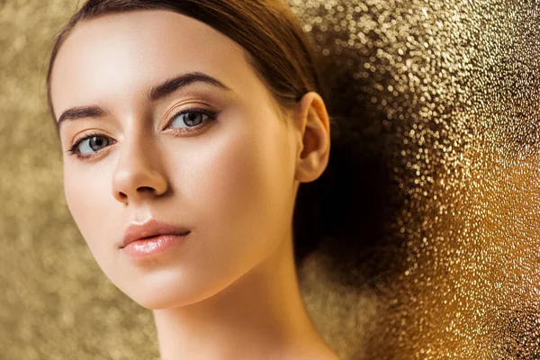 Молодая красивая женщина с блестящим макияжем смотрит в камеру на золотом текстурированном фоне — стоковое фото