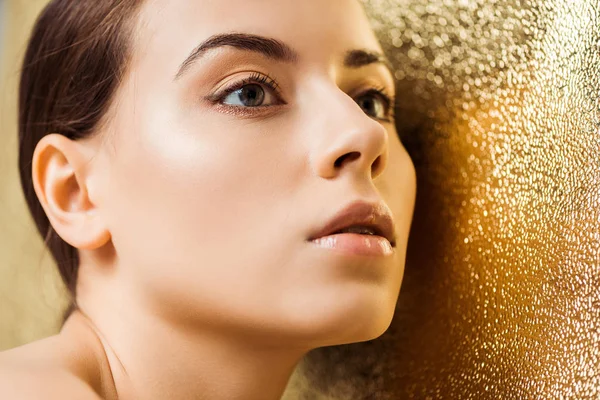 Mujer atractiva joven con maquillaje brillante mirando hacia otro lado sobre fondo de textura dorada - foto de stock