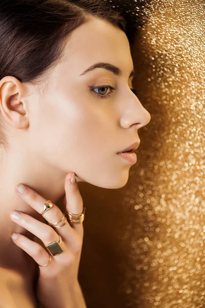 Mujer joven con maquillaje brillante y anillos de oro mirando hacia otro lado sobre fondo de textura dorada - foto de stock