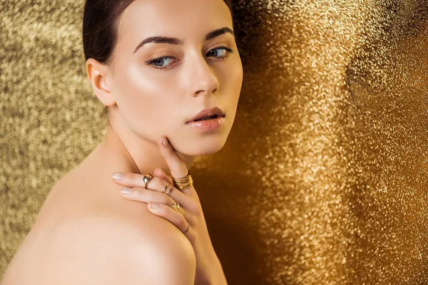 Mujer joven desnuda con maquillaje brillante y anillos de oro mirando hacia otro lado sobre fondo de textura dorada - foto de stock