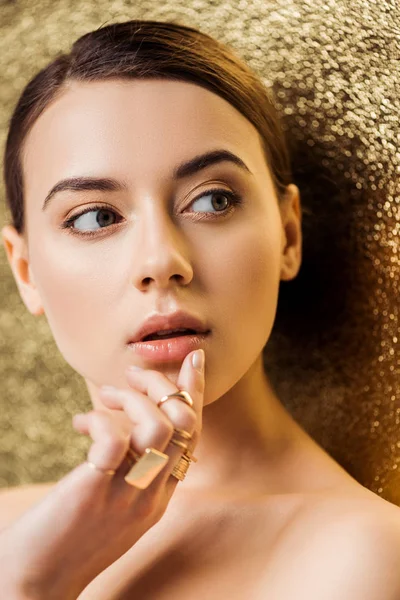 Mujer joven atractiva desnuda con maquillaje brillante y anillos de oro tocando los labios sobre fondo de textura dorada - foto de stock