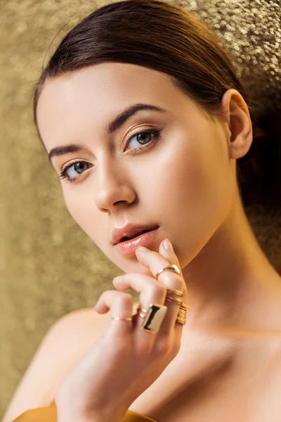 Attraktive junge Frau mit glänzendem Make-up und goldenen Ringen, die die Lippen auf goldenem strukturiertem Hintergrund berühren — Stockfoto