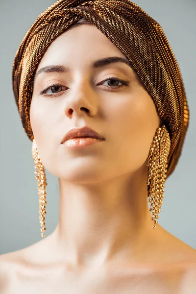 Молодая голая женщина с блестящим макияжем, золотые кольца в тюрбане глядя на камеру, изолированную на сером — стоковое фото