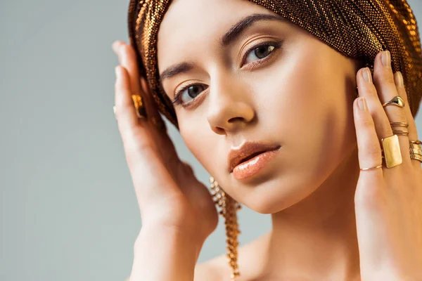 Jovem mulher nua com maquiagem brilhante, anéis dourados e brincos em turbante olhando para a câmera isolada em cinza — Fotografia de Stock